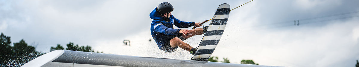 A person wearing a helmet and wetsuit performs a trick on a wakeboard, catching air above the water with a spray of droplets, against a cloudy sky background.