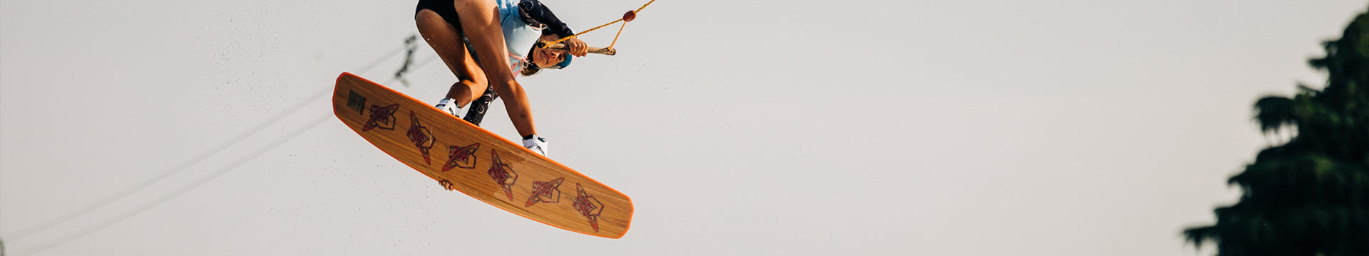 A person in mid-air wakeboarding, holding a rope handle, with bent knees and a large wooden board against a clear sky background.