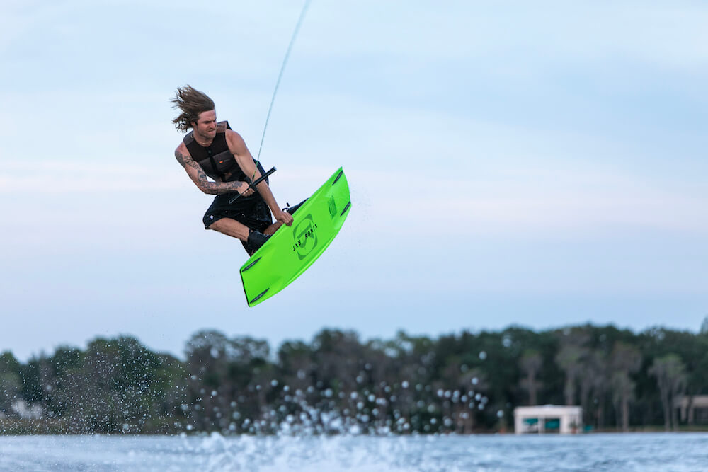 A long-haired rider jumps above the water while wakeboarding on the Ronix 2024 RXT Blackout Wakeboard, which features Blackout Technology for ultra-soft landings. They hold a tow rope, with trees and a house in the background.