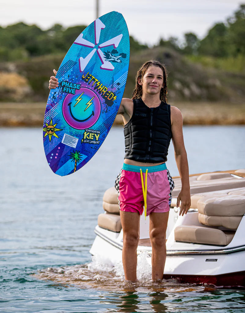 A woman standing on a boat holding a Phase 5 2024 Key Jett Shreds Wakesurf Board, with a Single Fin Setup.