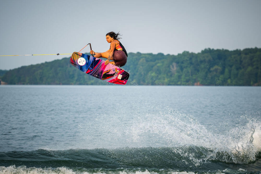 A woman showcasing her impressive wake boarding skills in the air above a serene lake, effortlessly gliding through the sky supported by the Hyperlite 2024 Prizm Wakeboard and experiencing heightened clarity with Hyperlite.