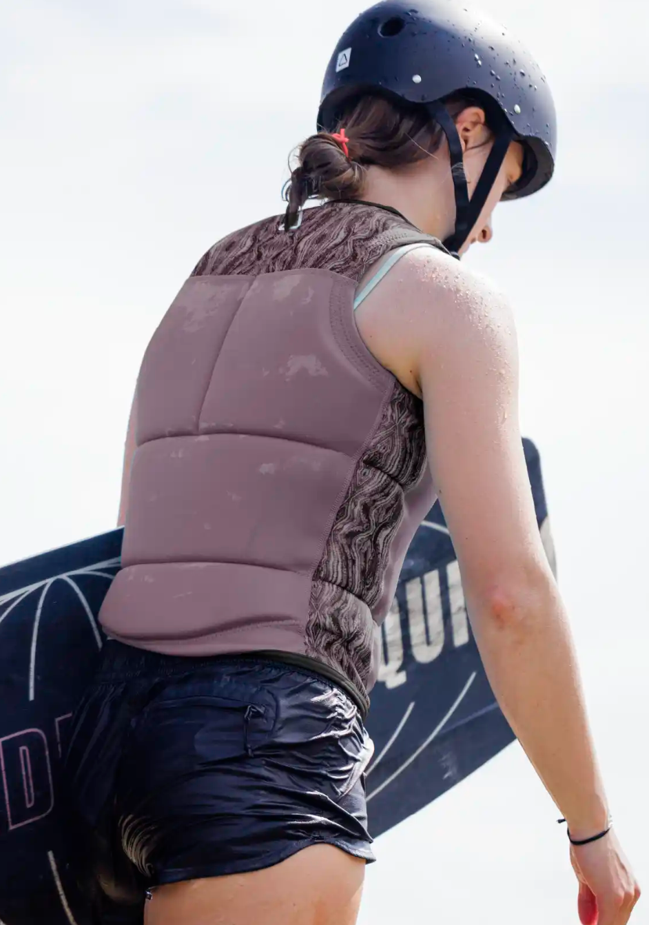 Wearing a Follow Harmony Ladies Jacket - Orchid by Follow Wake, a person in a helmet and black shorts holds a wakeboard outdoors, facing away from the camera.