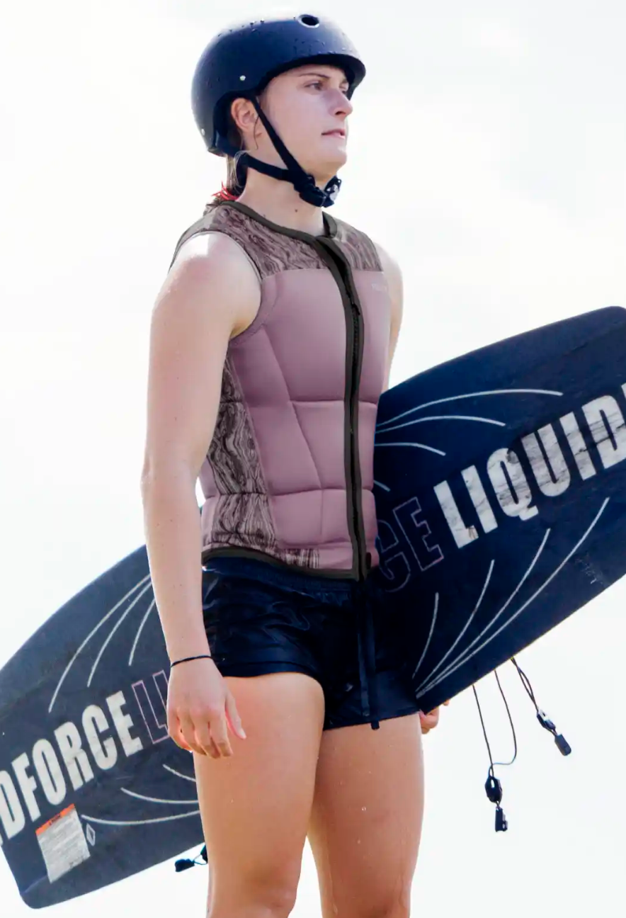 Wearing a Follow Harmony Ladies Jacket - Orchid by Follow Wake, a person in a helmet and black shorts stands holding a LIQUID FORCE wakeboard, gazing into the distance.