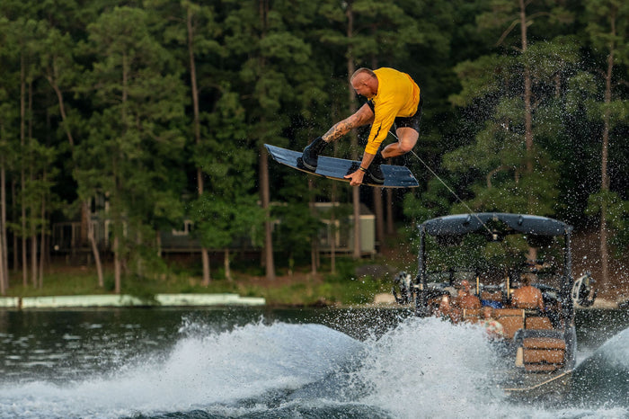 A man in a yellow shirt is doing a trick on a wakeboard, utilizing the Hyperlite 2023 Rusty Pro Wakeboard with Ultra Bindings.