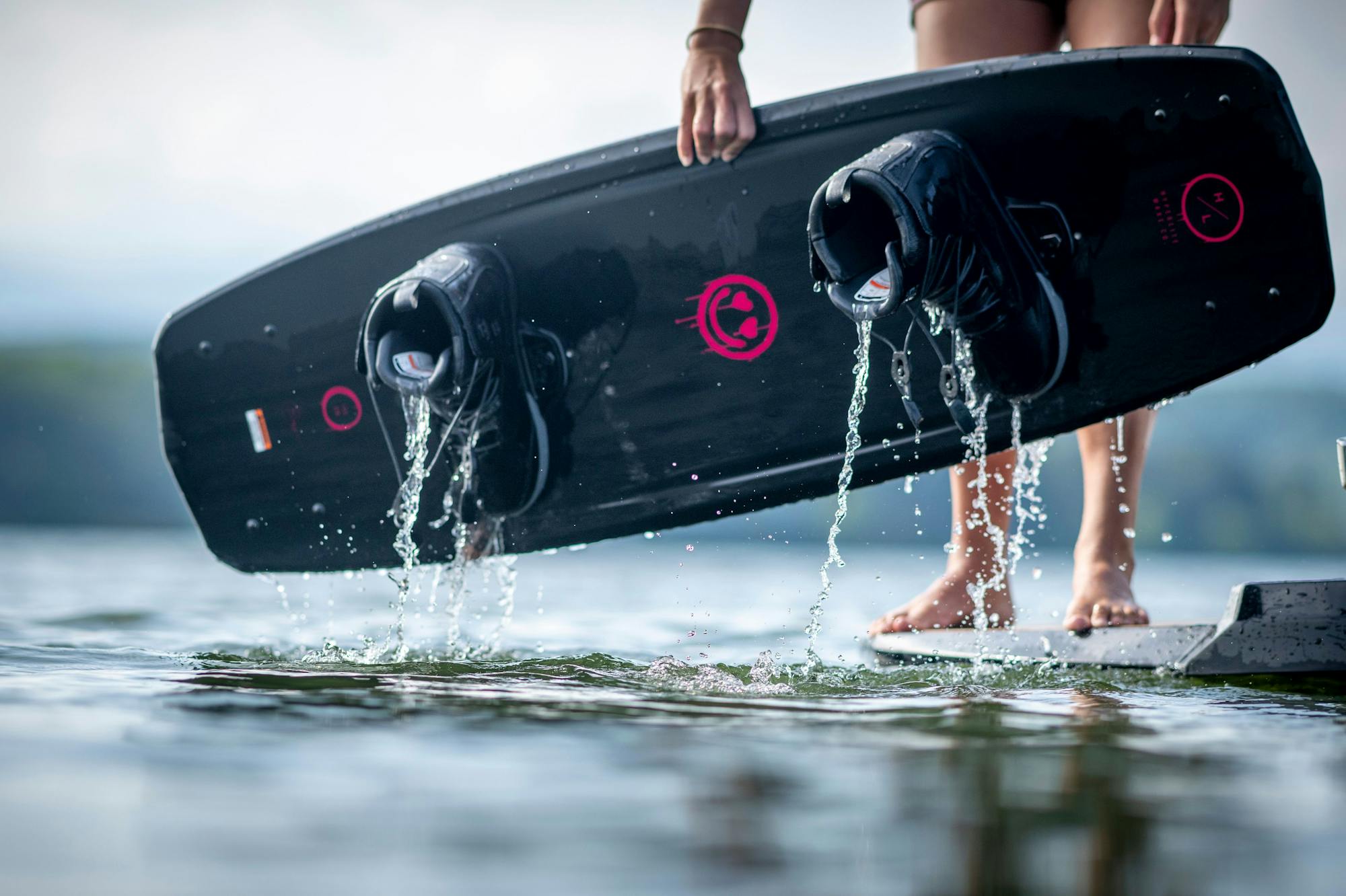 A person stands at the water's edge, holding their Hyperlite 2025 Journey Wakeboard with water draining from its boots. The board's 3-stage rocker is perfect for their next thrilling wakeboarding adventure.