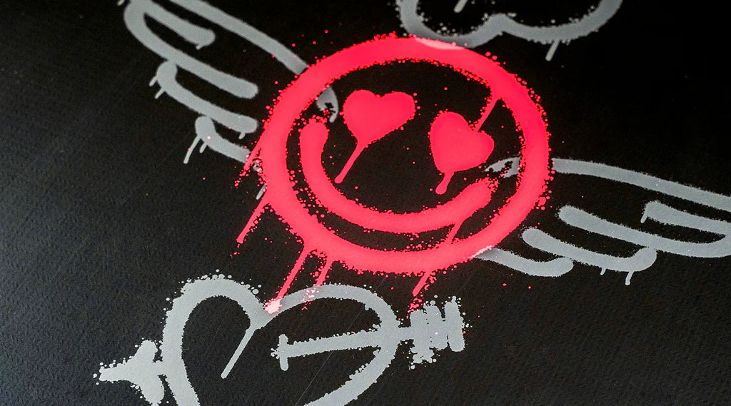 Set against a black backdrop, a spray-painted pink smiley face with heart eyes is highlighted by gray wings, capturing the exhilarating essence of wakeboarding. An arrow punctuates the design, evoking the lively spirit of Hyperlite's 2025 Journey Wakeboard with its 3-Stage Rocker in action.