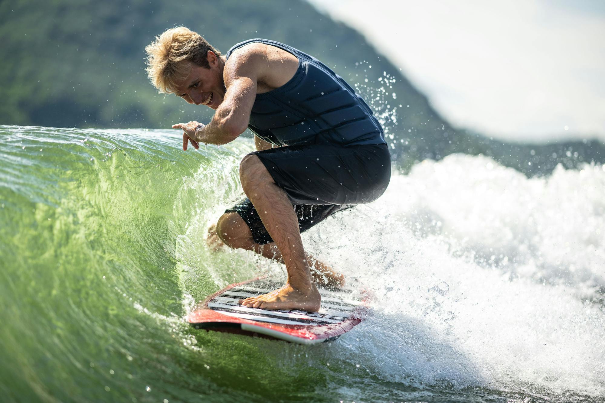 A man wakesurfing a wave in a sleeveless black wetsuit, using the Hyperlite 2025 Landlock Wakesurf Board - 5'9", with majestic mountains in the background.