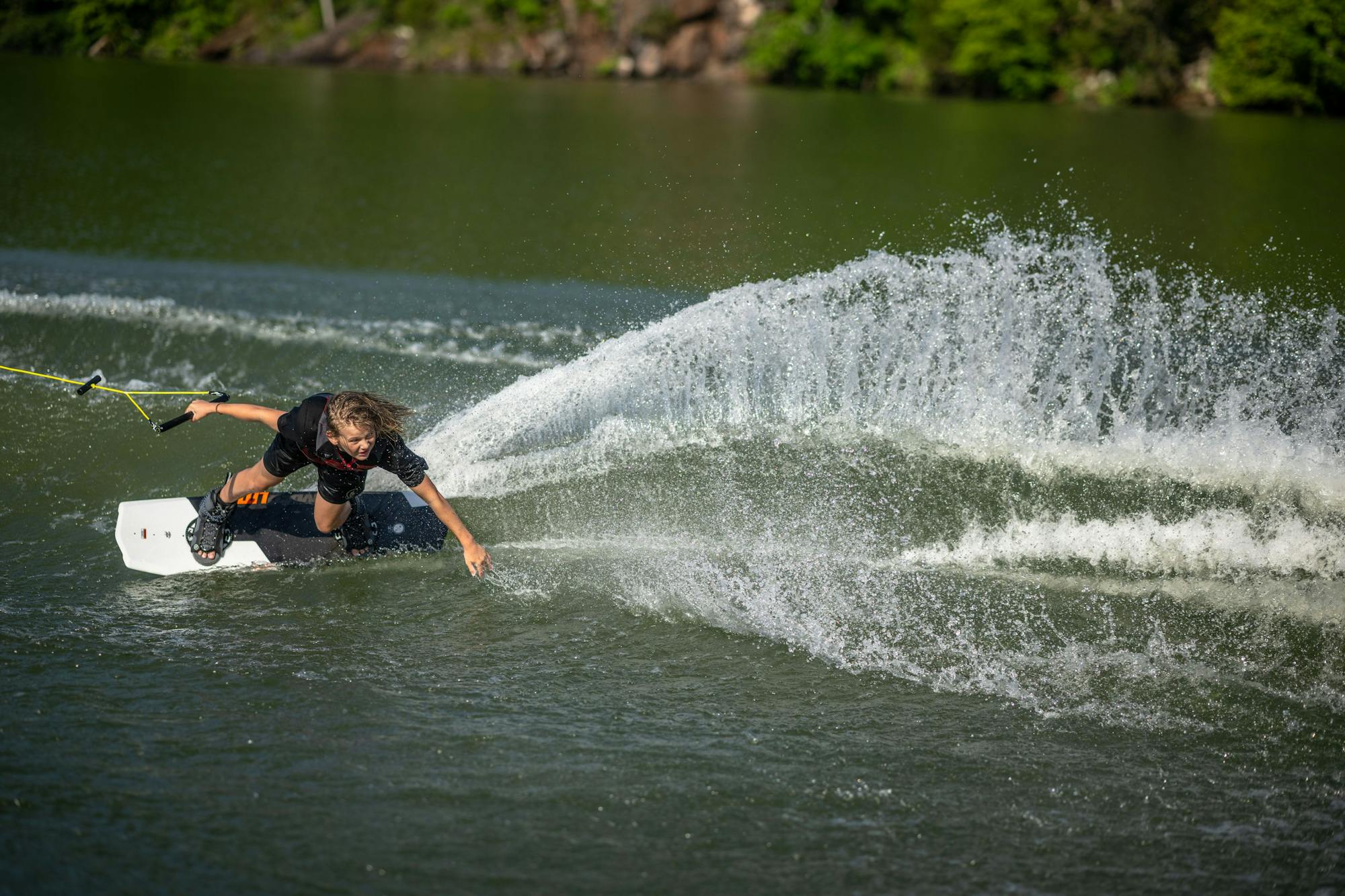 As a person wakeboards on the lake, they create a large spray of water while executing a sharp turn, highlighting the excitement of the Hyperlite 2025 State 2.0 Wakeboard design. Trees and rocks offer a picturesque backdrop as they skillfully carve through the waves.