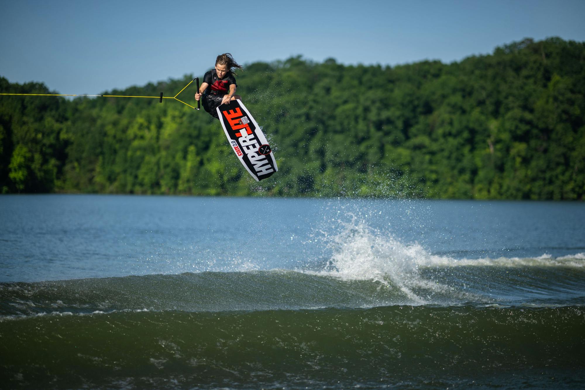 As a wakeboarder soars mid-air over the tranquil lake, they flaunt their skills using the Hyperlite 2025 State 2.0 Wakeboard against a forest-lined backdrop.