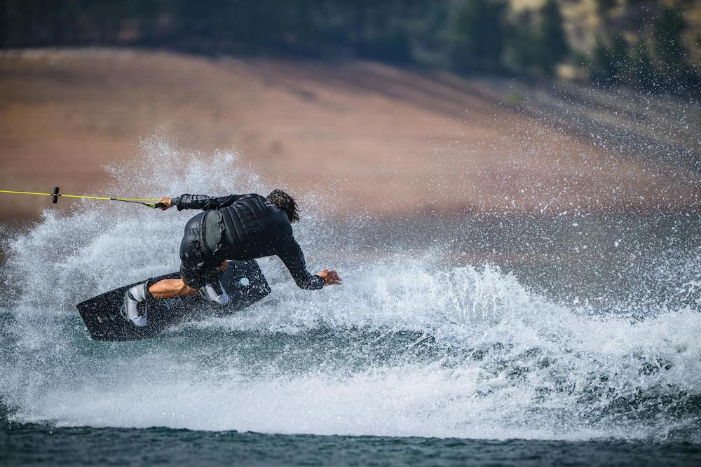 Wearing the ultra-lightweight Hyperlite Zero02 Boot 2026 by Hyperlite, a person in a black wetsuit wakeboards over water, leaning sharply and creating a large spray while holding a yellow tow rope with trees and land in the background.