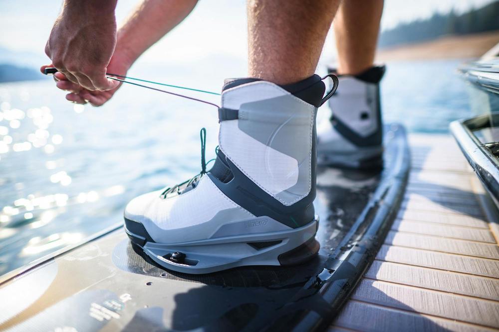 A person tightens the Hyperlite Zero02 Boot 2026 by Hyperlite while standing on a black wakeboard near the water, with blurred trees and shoreline in the background, highlighting the ultra-lightweight wake footwear.