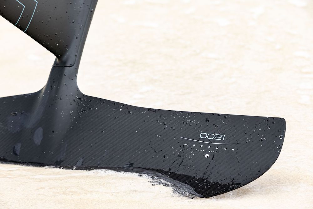 Close-up of a sleek black carbon fiber hydrofoil fin on a sandy surface, with water droplets glistening, showcasing the high-performance Liquid Force 2025 Carbon Horizon 1200 Fuselock Foil Set for ultimate stability.