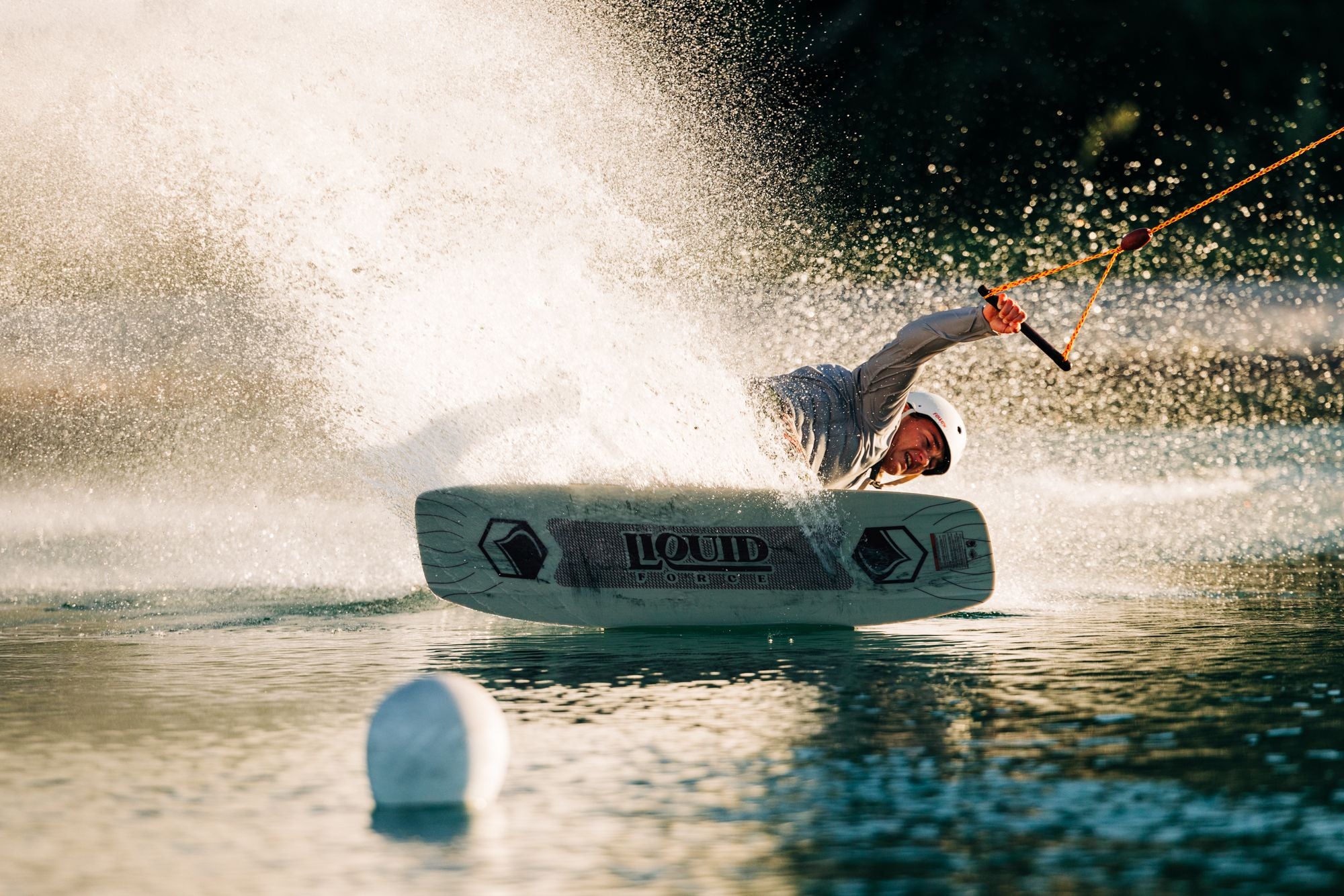 A person on the Liquid Force 2025 Noodle Wakeboard skillfully navigates over the water, flexing their body with precision while gripping a rope and generating an impressive splash.