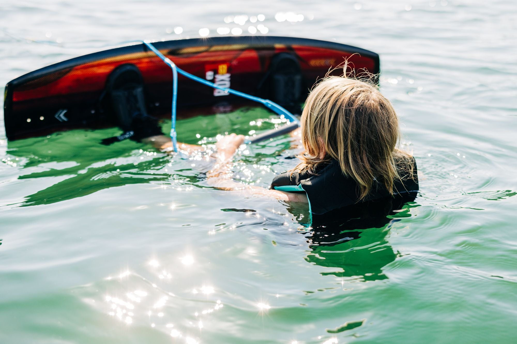 A young rider in the water confidently grips a wakeboard line, firmly holding onto a tow rope while wearing a reliable life vest. This scene embodies the spirit of the Liquid Force 2025 RDX Kid's Wakeboard, crafted with emerging talents in mind.