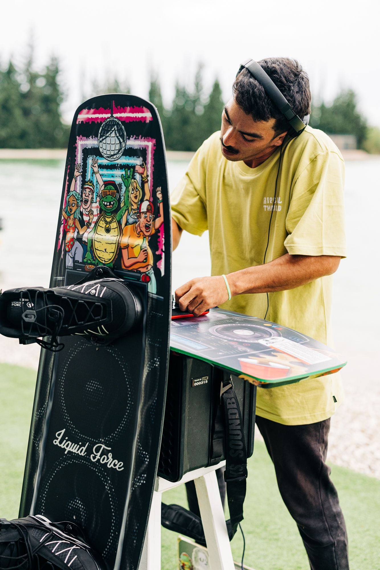 A person is adjusting DJ equipment on a stand outdoors, enhanced with Liquid Force technology, featuring a vibrant graphic display.