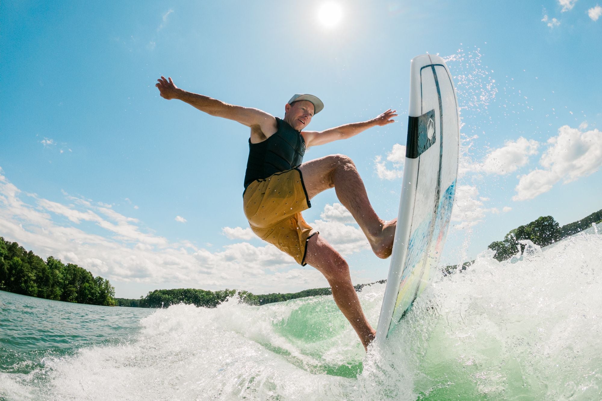 Gliding through the waves with arms outstretched, the surfer confidently displays a competition surf-style flair. Beneath a clear blue sky, equipped with a hat and life vest, they skillfully navigate past the green shoreline, propelled by the ultra-fast rocker profile of the Liquid Force 2025 Twin Turbo Wakesurf Board that enhances speed with every turn.