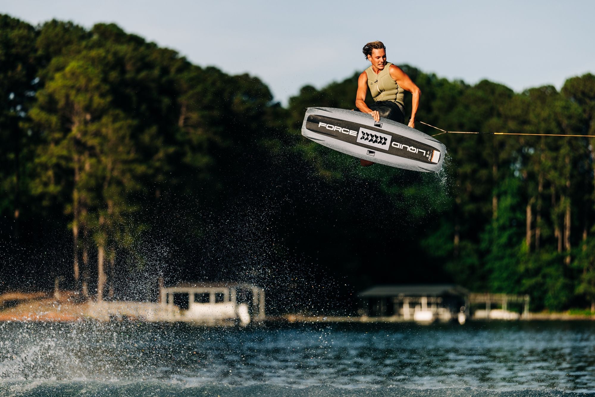 A person is wakeboarding in mid-air, gripping a rope, showing off their high-performance Liquid Force 2025 Unity Aero Wakeboard with CNC'd AERO Foam Core, against a stunning backdrop of trees and a serene lake.