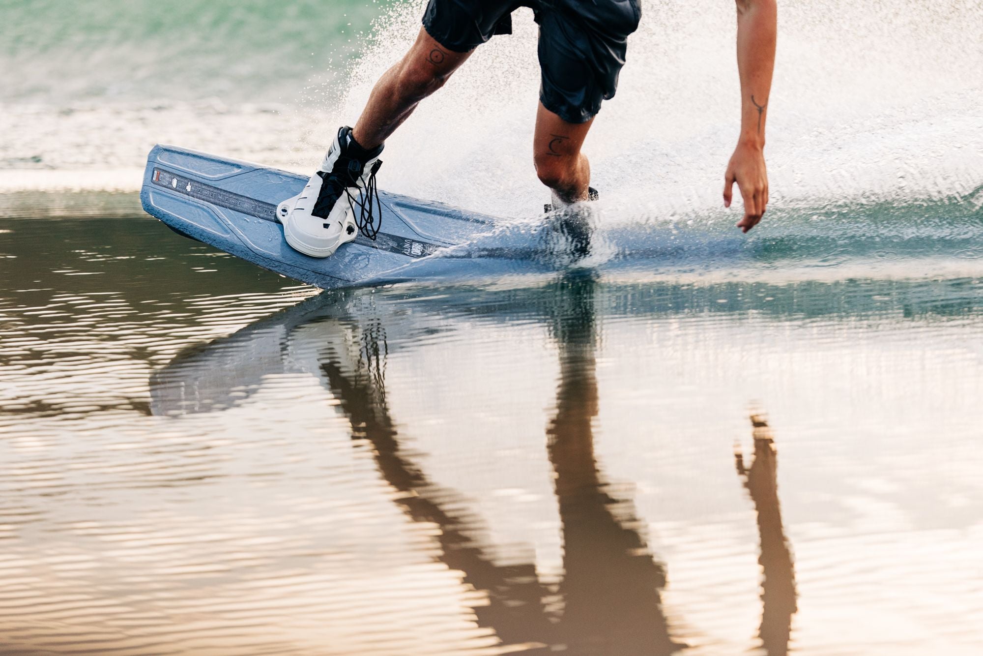 A person is wakeboarding on calm water, their high-performance Liquid Force 2025 Unity Aero Wakeboard gliding smoothly as they extend one arm and water splashes around. The board's CNC'd AERO Foam Core provides exceptional agility, and they're wearing shorts, fully immersed in the thrill of the ride.