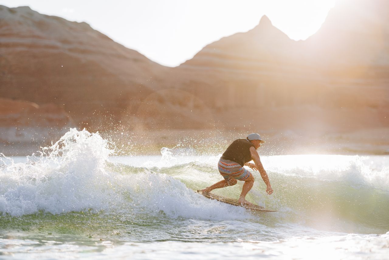 Wearing the Liquid Force Code Comp Vest 2026 - Black, a surfer rides a small wave in striped shorts and a cap, sunlight gleaming over rocky cliffs behind them.