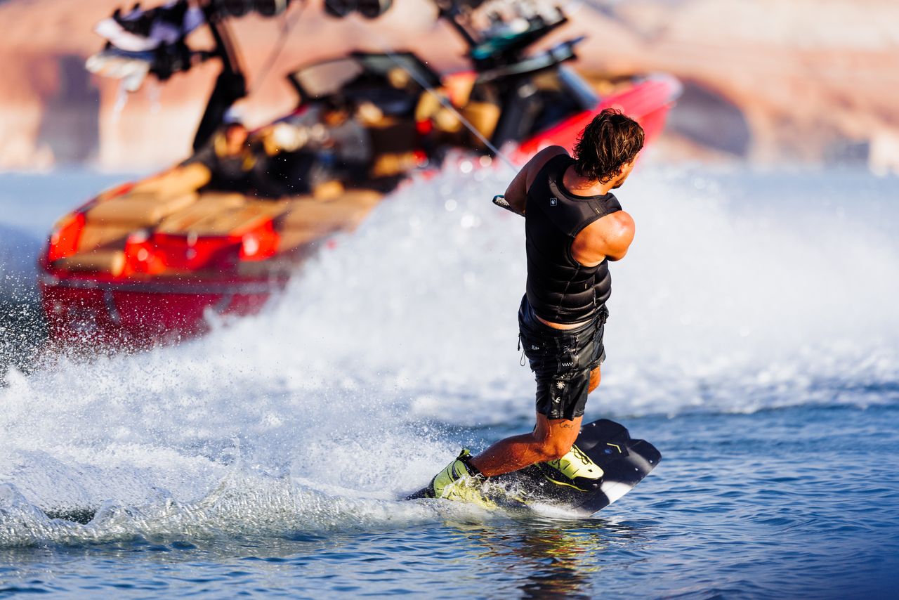 Riding on a lake, a wakeboarder sports the Liquid Force Code Comp Vest 2026 - Black while gripping a tow rope. A red boat carves waves in the sun, with rocky cliffs in the background. Brand: Liquid Force.