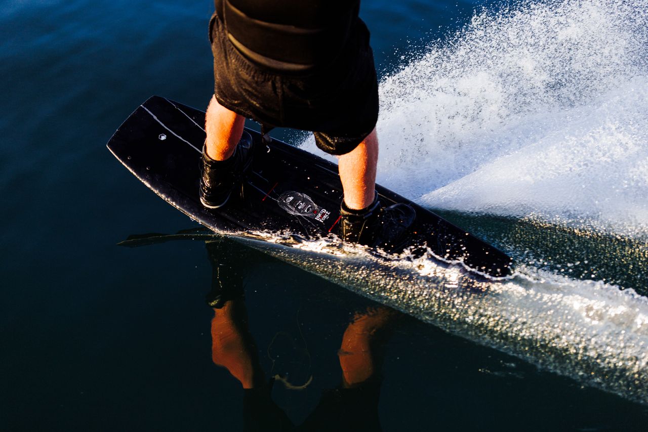 Close-up of a rider on the Liquid Force Origin Wakeboard 2026, water spraying behind as their feet are strapped in, wearing black shorts—capturing the excitement before BROstock 2025.