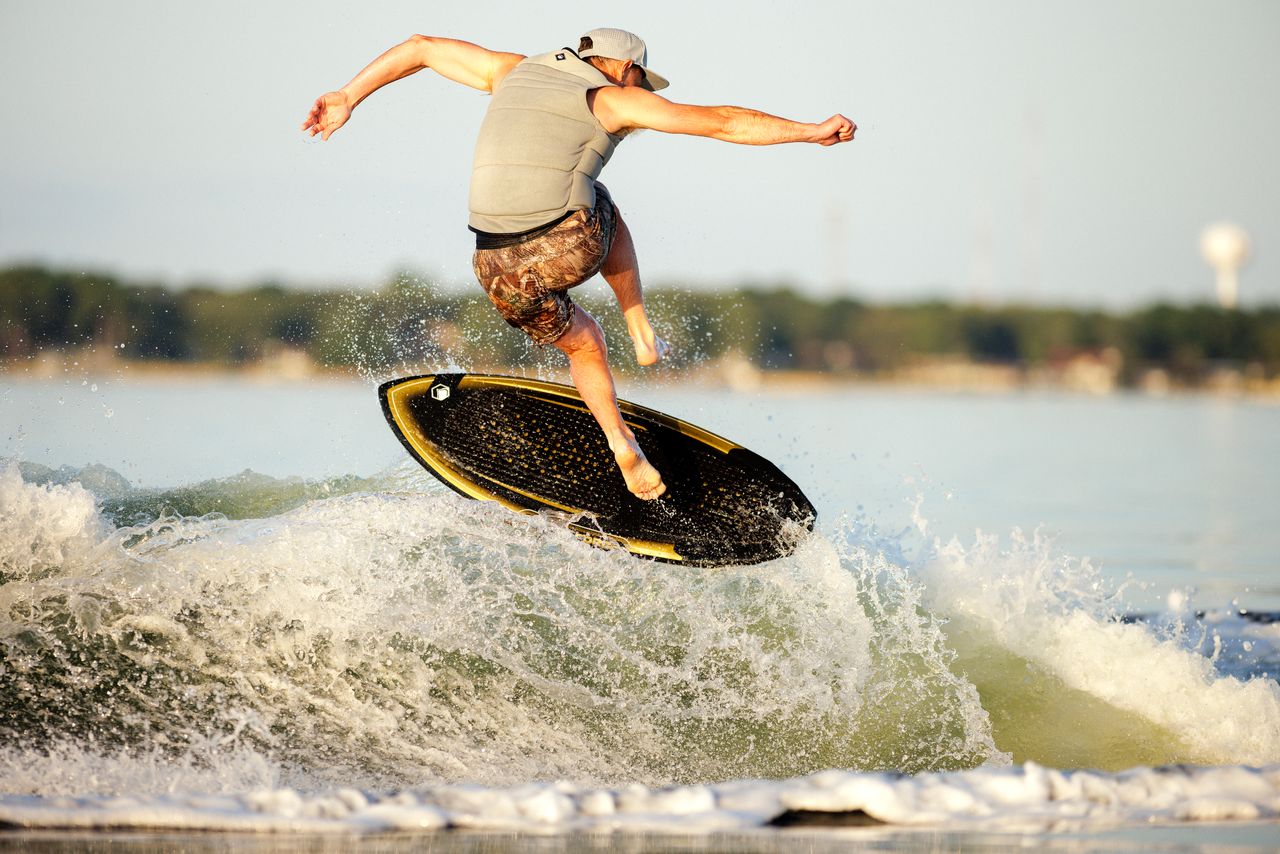 A person in a gray vest and cap performs a trick above a wave on a black and yellow Liquid Force Primo Wakesurf Board 2026, with water splashing and the background landscape blurred.