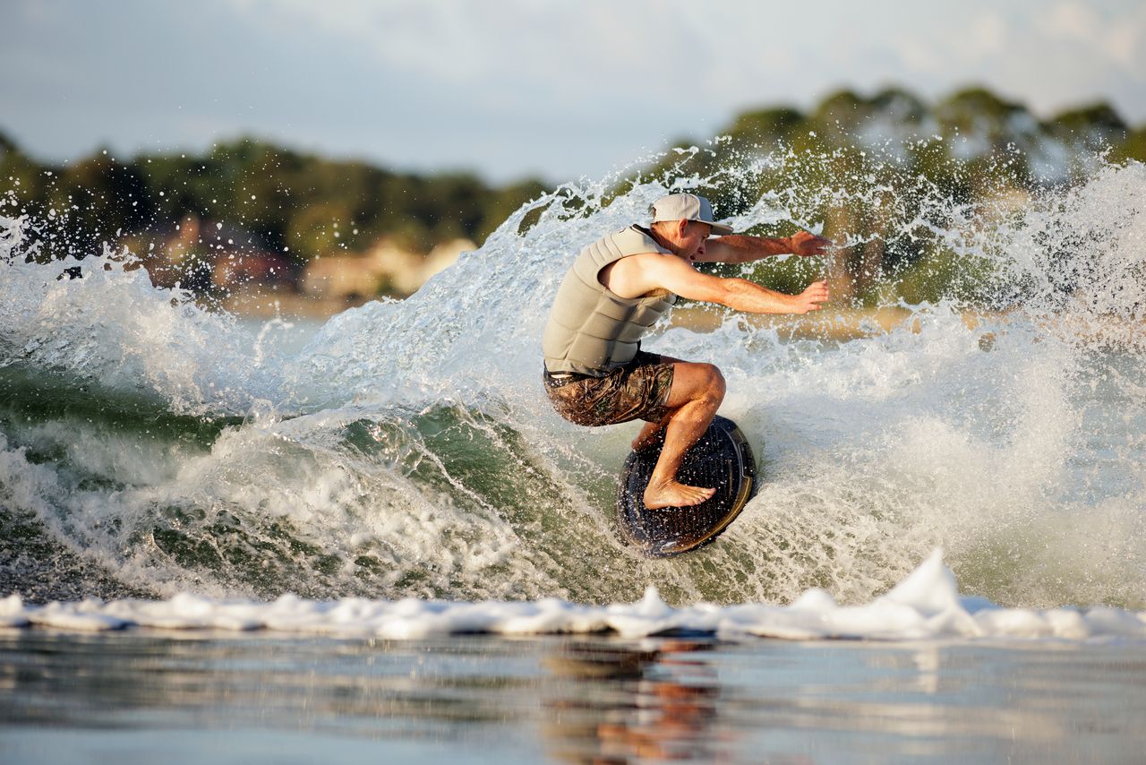 A person wearing a gray vest and cap rides the Liquid Force Primo Wakesurf Board 2026, carving through a wave and splashing water with trees and a blurred shoreline in the background.