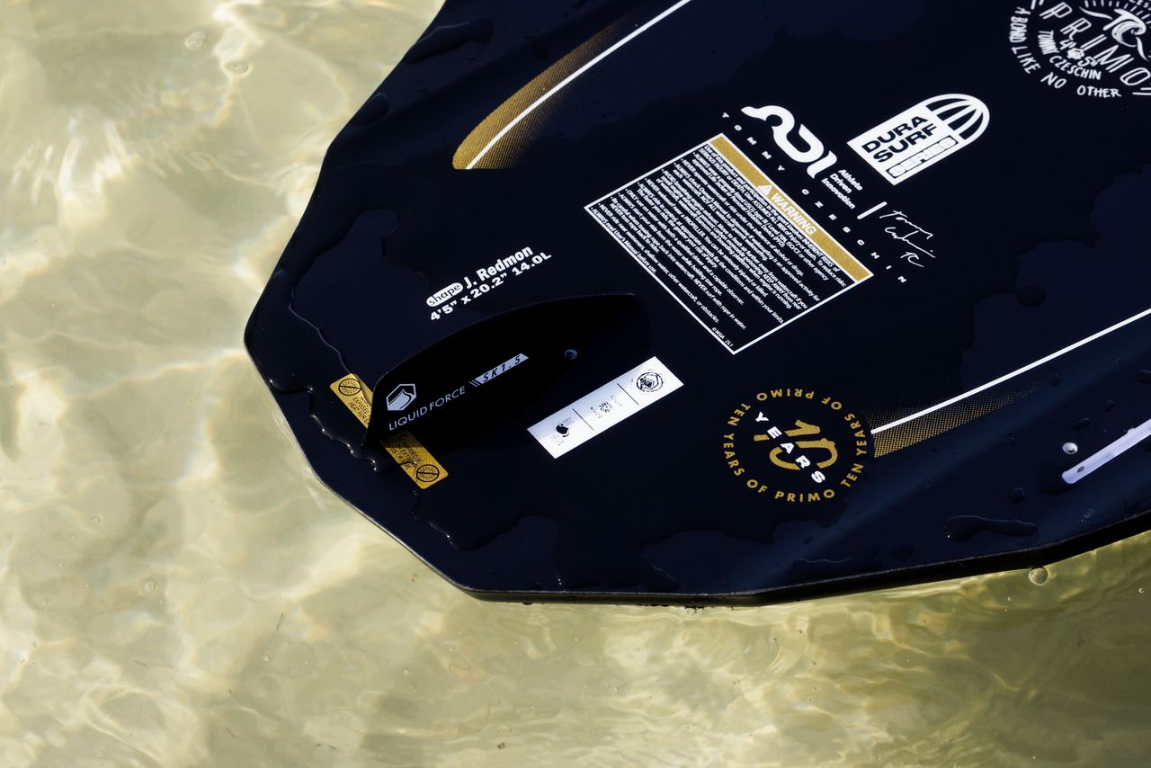 A close-up of the tail of the Liquid Force Primo Wakesurf Board 2026 shows branding and fin box details as it sits partially submerged in clear, shallow water. Various labels and Liquid Force logos are visible on the black board.