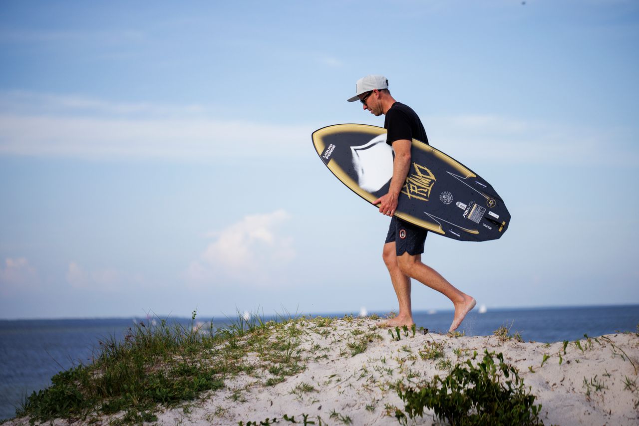 A man in a cap and black T-shirt walks barefoot on sandy grass, carrying the Liquid Force Primo Wakesurf Board 2026, with the ocean and blue sky in the background.