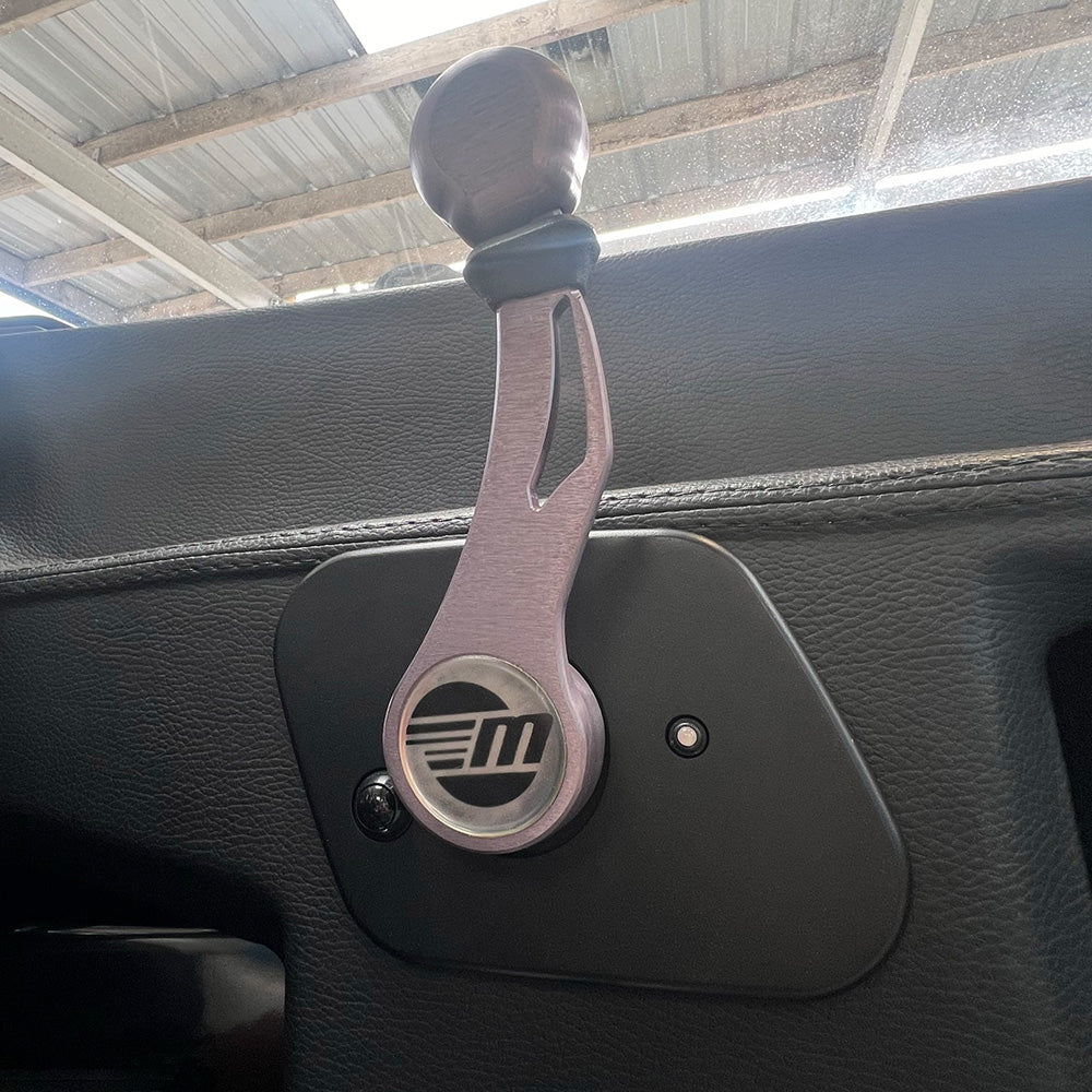 A close-up of the Malibu Shifter '15-'19 by Malibu features its unique metallic design and M logo, mounted on a black car door panel under a corrugated metal roof, echoing the style used in 2015-2019 Malibu boats.