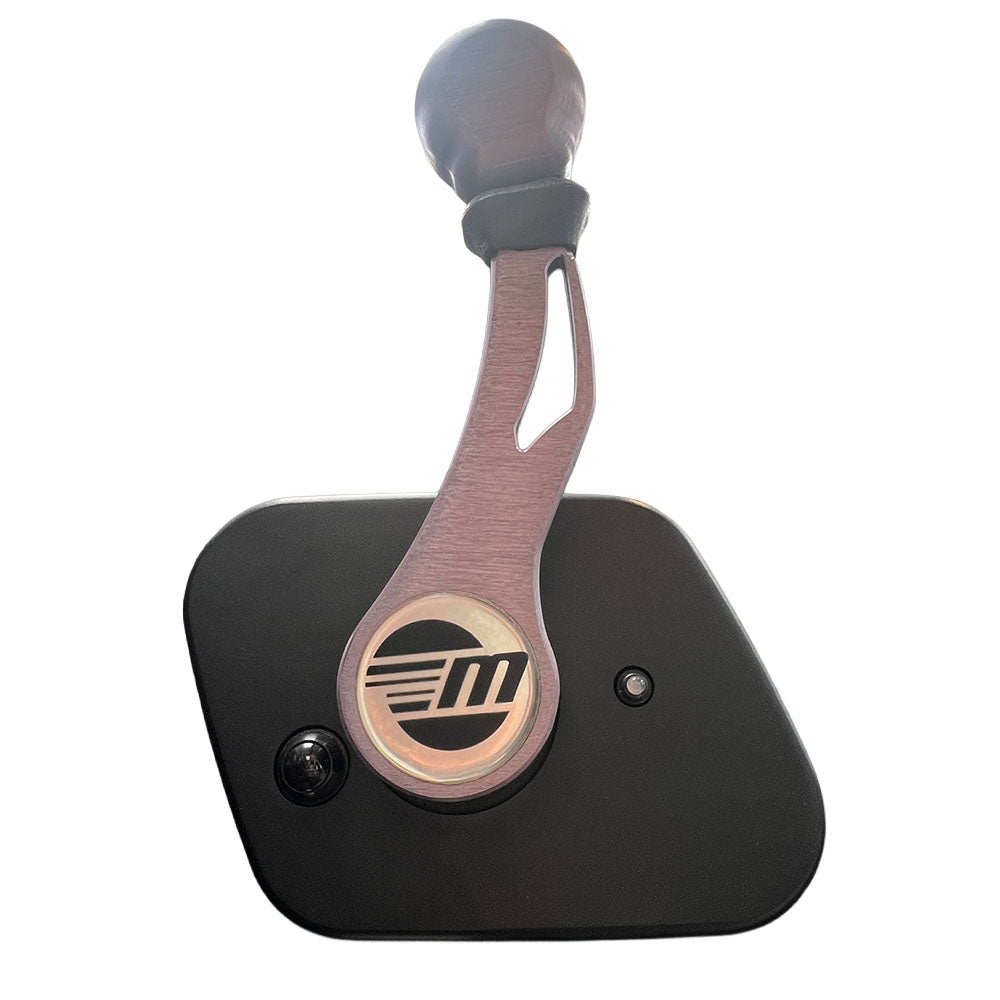 A close-up of the Malibu Shifter '15-'19 by Malibu, featuring a metallic-finish handle with a stylized “M” logo on a black door panel. Sunlight and a metal roof evoke classic Malibu boat designs from 2015-2019.