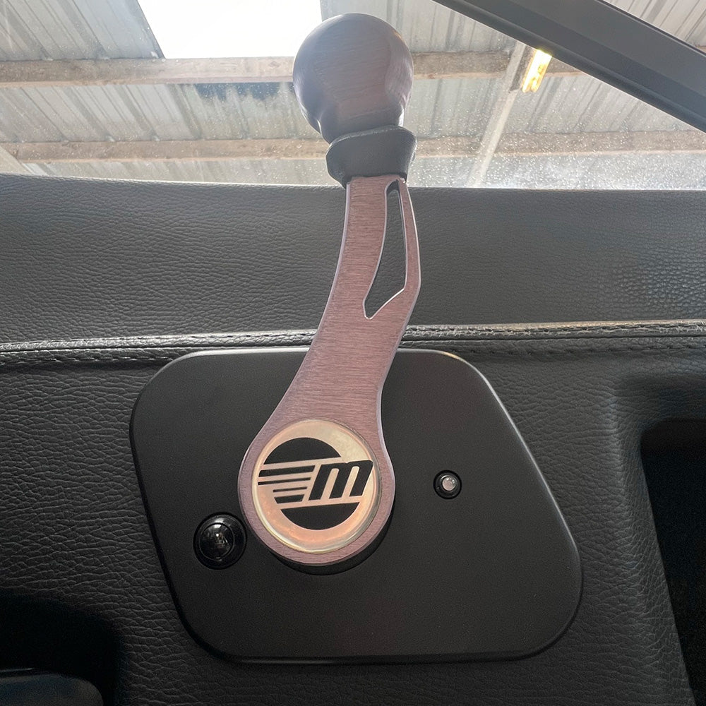 A close-up of the Malibu Shifter '15-'19 by Malibu, featuring a metallic-finish handle with a stylized “M” logo on a black door panel. Sunlight and a metal roof evoke classic Malibu boat designs from 2015-2019.