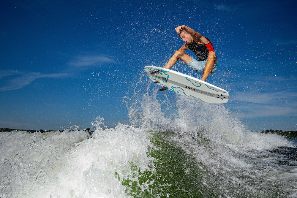 Parker Payne, wearing a red and black shirt, performs an impressive aerial trick on a wakeboard above splashing water. On this sunny day, he demonstrates his skills with agility reminiscent of the Phase 5 2025 Phantom Wakesurf Board.