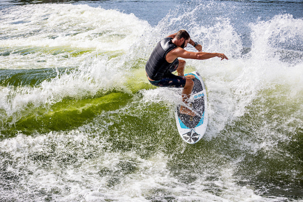 A man in a black life vest surfs a wave on the Phase 5 Ahi Wakesurf Board 2026, balancing as water splashes around him beneath a bright sky.