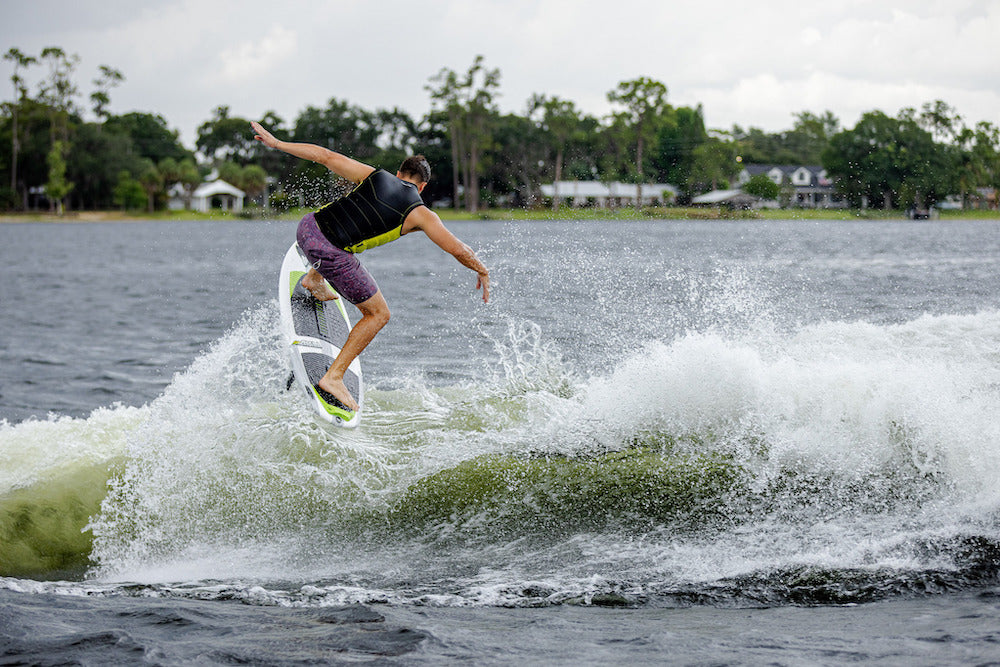 A person in a life vest and shorts wakesurfs on a lake, performing a trick on their 2026 Phase 5 Aku v2 Wakesurf Board, with trees and houses in the background.