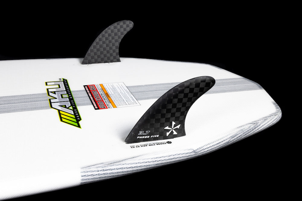 Close-up of the tail of a white Phase 5 Aku v2 Wakesurf Board 2026 with two black checkered fins, various decals, and labels on a black background.