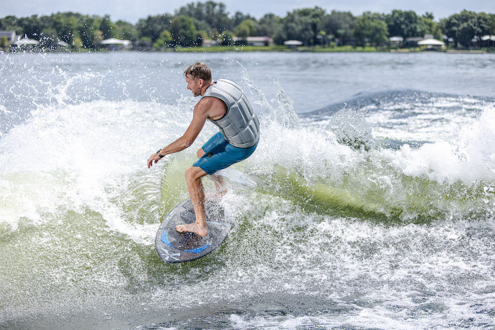 A man in a life vest and blue shorts rides a Phase 5 Diamond Turbo Wakesurf Board 2026, balancing with bent knees and outstretched arms as trees and houses appear in the background.