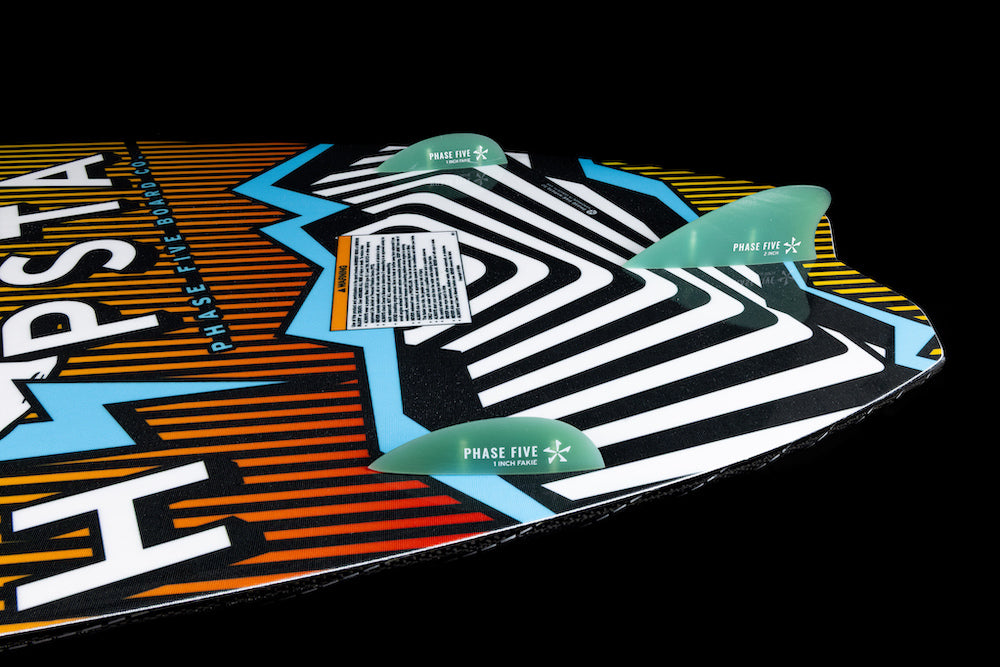 Close-up of the Phase 5 Hypsta v2 Wakesurf Board 2026 tail, featuring bold geometric patterns in black, white, yellow, blue, and orange, a TRI-Fin setup with three green Phase 5 fins, and a warning label on the surface.