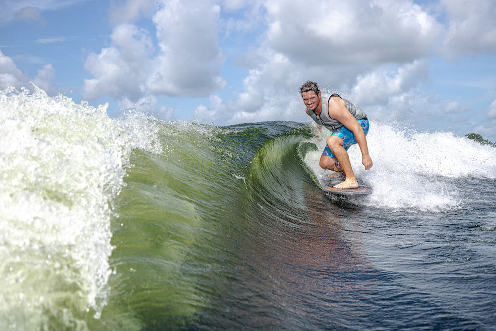 A man in blue shorts rides a wave on a Phase 5 Model X Wakesurf Board 2026, smiling and crouched low as he surfs near the wave's crest under a partly cloudy sky.