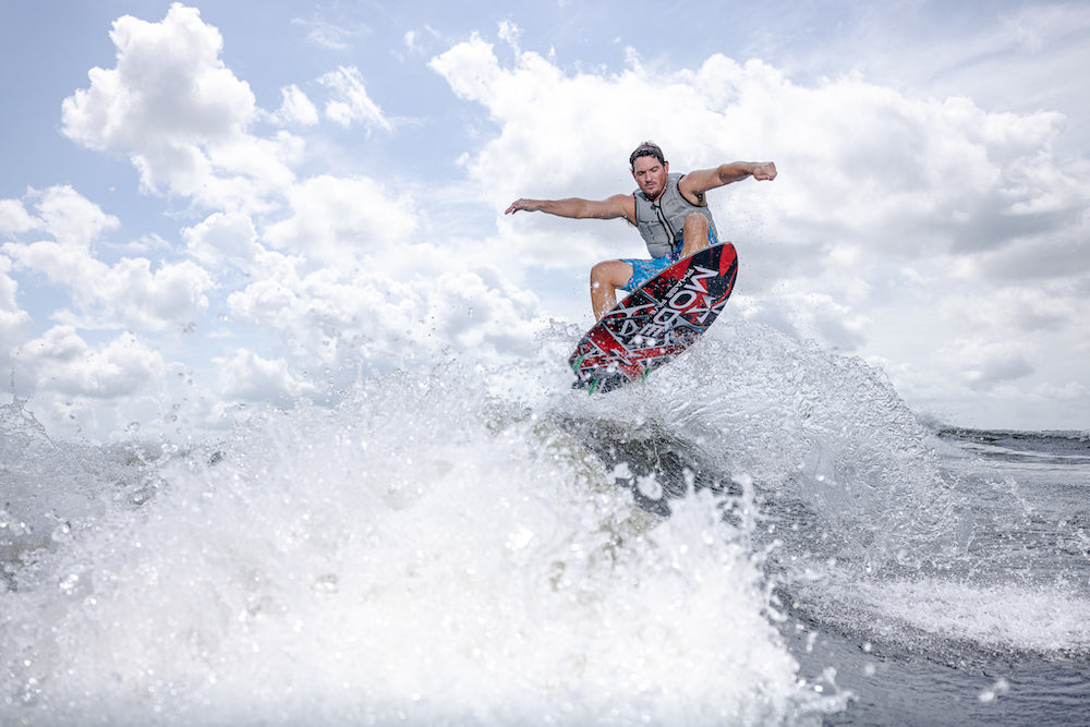 A person in a life vest is airborne on a Phase 5 Model X Wakesurf Board 2026 above splashing water, with a blue sky and clouds in the background.