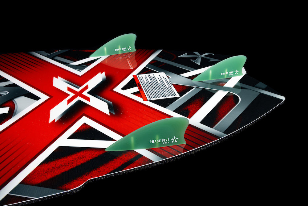 Close-up of the Phase 5 Model X Wakesurf Board 2026, featuring a bold red, black, and white X design with three green fins and a GatorSkin glass finish. The Phase 5 brand and a white warning label are also visible on the board.