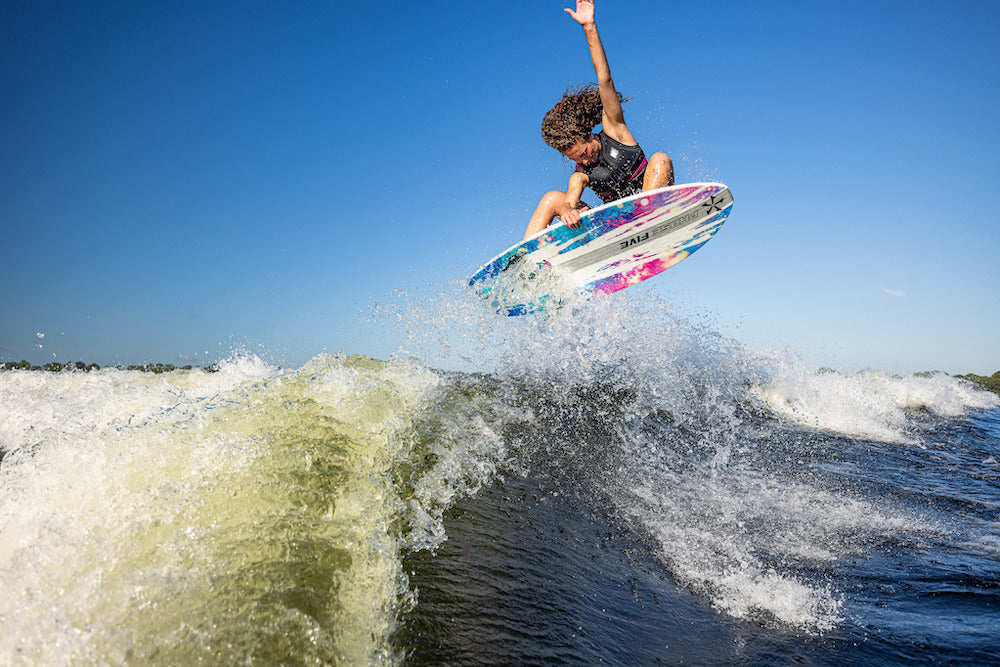 A curly-haired wakesurfer jumps high above the wake on a sunny day, riding the lightweight Phase 5 Rush Wakesurf Board 2026, with water spray and blue skies showcasing its performance.