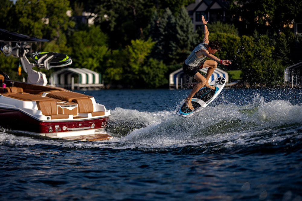 Riding a Phase 5 Swell Wakesurf Board 2026, a person does a trick on the wake behind a boat on a lake, with trees and lakeside houses visible in the background.
