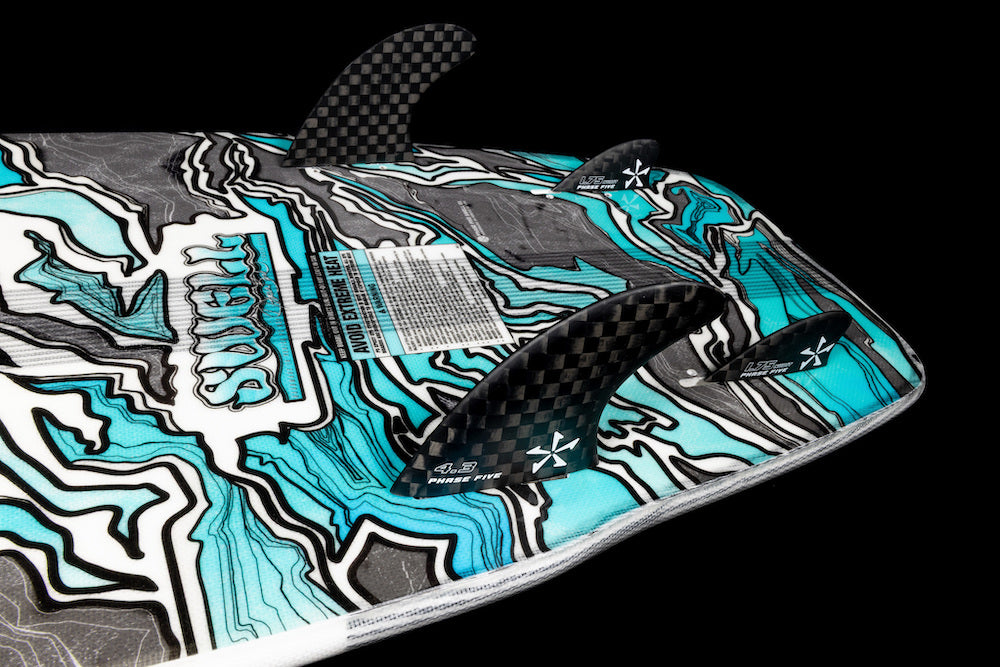 Close-up of the underside of the 2026 Phase 5 Swell Wakesurf Board, featuring a blue, black, and white abstract pattern and three black carbon fiber fins, set against a black background. A bold surf-style board from Phase 5.