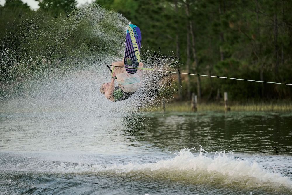 A wakeboarder on the Radar Graviton Trick Waterski 2026 by Radar nails a backflip with a tow rope, landing smoothly in a spray of water, waves up front, and green trees framing the scene.