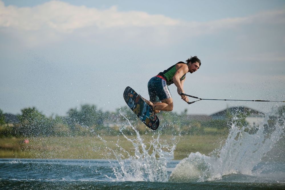 A person rides the Radar Graviton Trick Waterski 2026 by Radar, catching air above the lake while holding a tow rope, performing stylish trick skiing moves with smooth landings and splashes against a grassy shoreline.