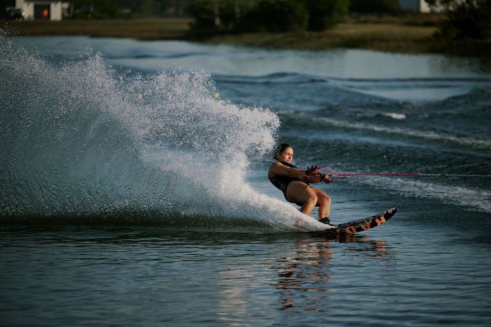 A woman on the Radar Lyric Women's Waterski | Lyric Boa + Boa ARTP 2026 glides across the lake, carving the water and sending up a spray, with trees and grassy land in the background.
