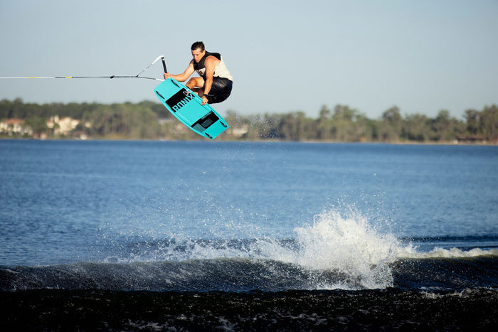 A person riding on the Ronix 2025 District Wakeboard paired with District Bindings, performing explosive jumps and executing a stunning aerial trick over the water, set against a serene lake backdrop.