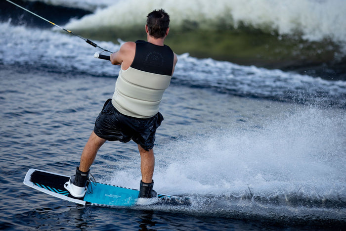 A person expertly wakeboarding on a body of water, equipped with the Ronix 2025 District Wakeboard and District Bindings, dons a life vest and grips a tow rope as they perform explosive jumps against the backdrop of waves, showcasing the thrill of gliding with the hybrid rocker design from Ronix.