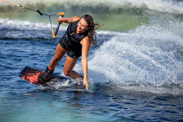 A person expertly wakeboarding on a lake with the Ronix 2025 Krush Wakeboard | Divide Bindings, creating a splash as their hand skims the water. They sport a life jacket while mastering the waves in the background.